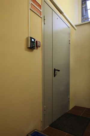 Дверь в серверную. На стене расположена биометрическая система доступа в серверную, работающая по отпечаткам пальцев. На полу рядом с дверью находится аппарат для автоматического одевания бахил (для рабочего персонала и посетителей серверной).
